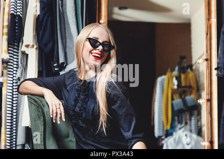 Mode blond lächelnde Mädchen in schwarzen Kleid tragen Sonnenbrillen sitzen auf dem grünen Sessel im Einkaufszentrum, schwarzer Freitag Konzept Stockfoto