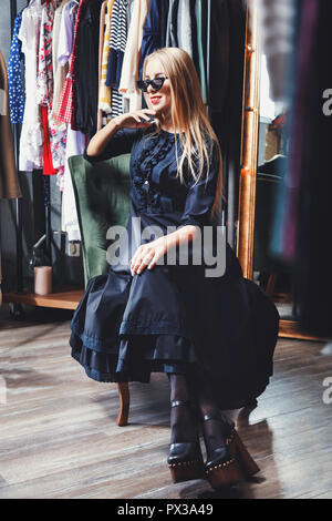 Mode blond lächelnde Mädchen in schwarzen Kleid tragen Sonnenbrillen sitzen auf dem grünen Sessel und schauen auf das Fenster im Einkaufszentrum, schwarzer Freitag Konzept Stockfoto