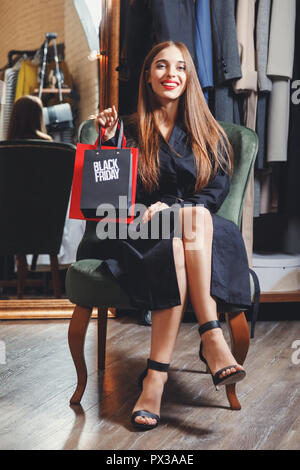 Schwarzer Freitag elegante reiche Frau tragen modische Trench-Mäntel im Sessel sitzend mit Einkaufstaschen in Fashion Mall nach groß einkaufen, Konzept des Konsumismus, Verkauf, reiches Leben Stockfoto