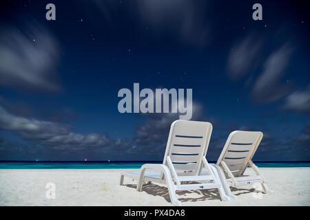 Zwei leere Liegestühle stehen auf dem Ozean Sand Strand unter Sternenhimmel mit Stars und ziehenden Wolken. Lange Belichtung geschossen mit weiten Blickwinkel Stockfoto