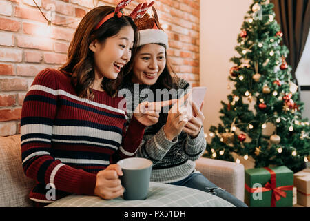 Beste Freunde mit Weihnachtsmütze und Rehe sind online einkaufen zu Hause mit Smartphone. Große Weihnachten Verkauf im Dezember. überrascht, der auf die erstaunliche Pr Stockfoto