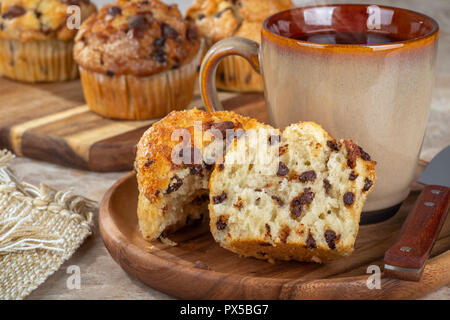 Nahaufnahme eines Chocolate Chip muffin halbieren und eine Tasse Kaffee auf einer Holzplatte mit Muffins im Hintergrund Stockfoto