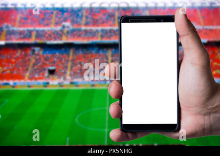 Mans hand mit einem Smartphone gegen Fußball oder das Fußballstadion Hintergrund. Weiße leere Bildschirm Bereit für mockup Präsentation. Stockfoto