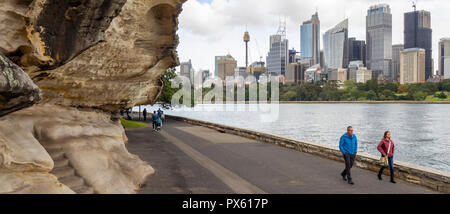 Fußgänger auf dem Weg entlang Farm Cove Sydney Harbour mit Bürogebäude und CBD Skyline an einem bewölkten Tag Sydney, NSW, Australien. Stockfoto