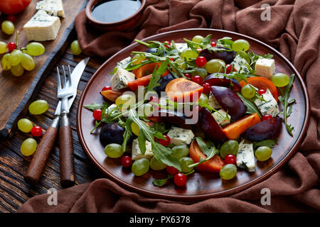 In der Nähe von köstlichen Herbst Salat mit Schokolade Kaki, Blauschimmelkäse, grüne Trauben, Pflaumen- schichten und Rucola auf einem Steingut Teller mit Zutat Stockfoto