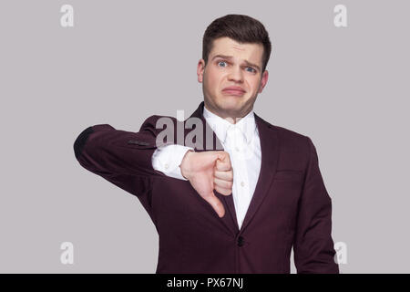 Portrait von Stattlichen verwirrten jungen Mann in violett Anzug und weißes Hemd, stehen, zu Kamera suchen mit Daumen nach unten und unbefriedigt. indoor Stud Stockfoto