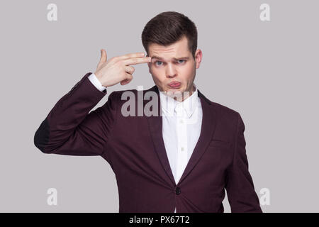 Portrait von verwirrt hübscher junger Mann in violett Anzug und weißes Hemd, stehen, zu Kamera mit ernsten Gesicht und gun Geste. indoor Studio s Stockfoto