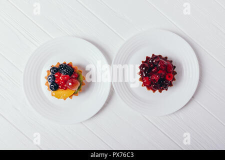 Kuchen mit Beeren auf weißem Tabelle, Ansicht von oben. Zwei Desserts mit Blaubeeren, Brombeeren, Johannisbeeren, Kirschen, Trauben auf Tellern serviert, flach. Stockfoto