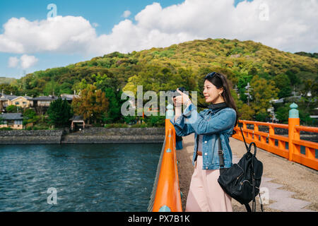 Elegante asiatischen reisenden unter Foto auf der Brücke mit schönen Berg im Hintergrund. Lady objektiv Mann fotografieren der saubere Fluss in Kyoto. spr Stockfoto