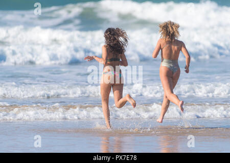 Zwei junge attraktive weibliche Urlauber, die in einem Aufenthalturlaub am Fistral in Newquay in Cornwall ins Meer laufen. Stockfoto