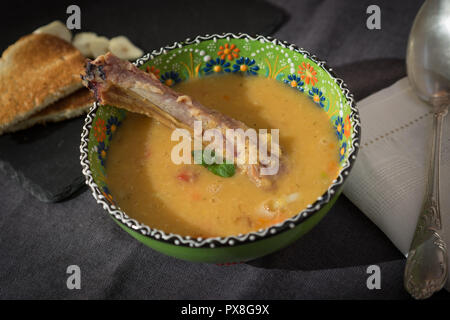 Rote Linsensuppe mit geräuchertem Rippchen in einem hellen Grün Schüssel mit floralen Ornament. Dunkelgrau Stoff Hintergrund. Stockfoto