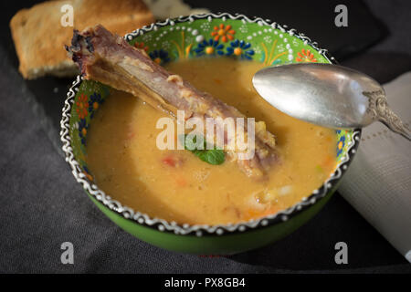 Rote Linsensuppe mit geräuchertem Rippchen in einem hellen Grün Schüssel mit floralen Ornament. Dunkelgrau Stoff Hintergrund. Stockfoto