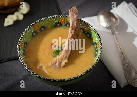 Rote Linsensuppe mit geräuchertem Rippchen in einem hellen Grün Schüssel mit floralen Ornament. Dunkelgrau Stoff Hintergrund. Stockfoto