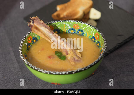 Rote Linsensuppe mit geräuchertem Rippchen in einem hellen Grün Schüssel mit floralen Ornament. Dunkelgrau Stoff Hintergrund. Stockfoto