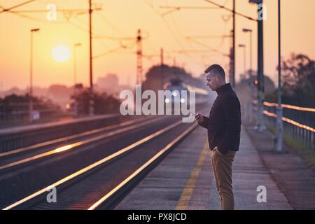 Junger Mann am Bahnhof Plattform warten und mit smart phone gegen Pendeln Zug bei Sonnenaufgang. Stockfoto