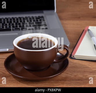 Nahaufnahme einer dampfenden Tasse Kaffee auf einem hölzernen Schreibtisch mit einem Laptop im Hintergrund Stockfoto