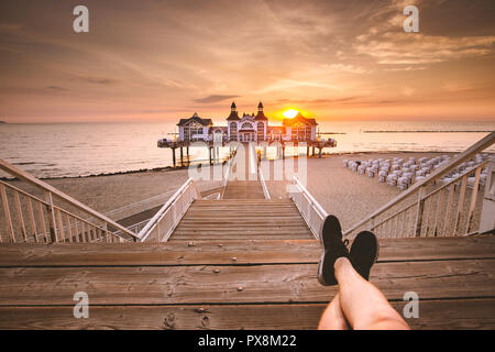 Junge Mann sitzt auf hölzernen Pier genießen Sie einen wunderschönen Sonnenaufgang am berühmten Seebrucke Sellin in Ostseebad Sellin, Insel Rügen, Ostsee, Deutschland Stockfoto