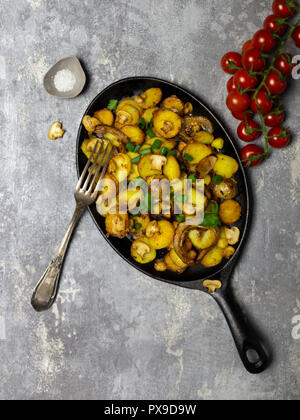 Rustikale frische Bratkartoffeln mit Pilzen und Zwiebeln in eiserne Pfanne mit Gabel und Salz an grauer Hintergrund Stockfoto