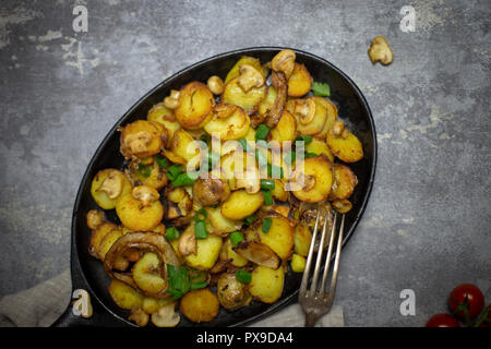 Rustikale frische Bratkartoffeln mit Pilzen und Zwiebeln in eiserne Pfanne mit Gabel auf grauem Hintergrund Stockfoto