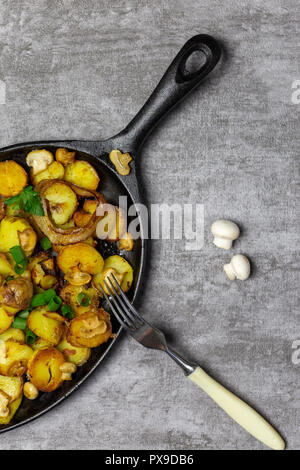 Traditionelle frische Bratkartoffeln mit Pilzen und Zwiebeln in eiserne Pfanne mit Gabel und frische Pilze auf grauem Hintergrund Stockfoto