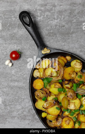 Traditionelle frische Bratkartoffeln mit Pilzen und Zwiebeln in eiserne Pfanne mit frischen Pilzen und Tomaten auf grauem Hintergrund Stockfoto