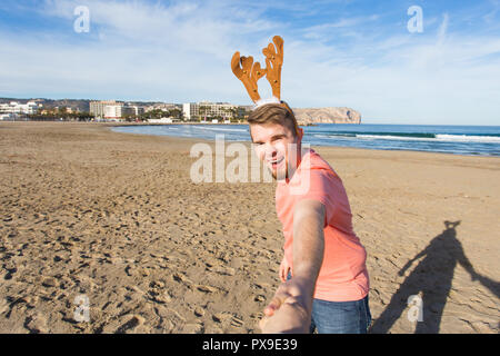 Folgen Sie mir, Ferien und Weihnachten Konzept-fröhlicher junger Mann mit Hirsch Hörner holding Santa's Hand am Strand. Stockfoto