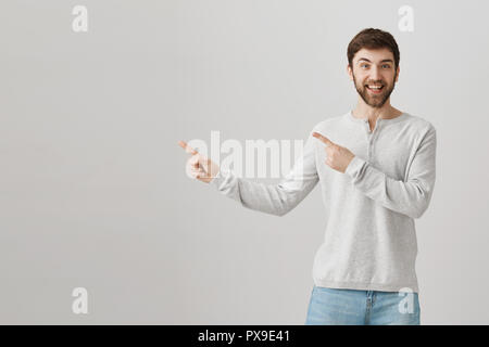 Gerne gut aussehenden Kaukasier bärtigen Kerl mit beiden Zeigefingern links, lächelnd und wird beim Stehen über grauer Hintergrund aufgeregt. Upbeat Freund um Erlaubnis bittet, zu Achterbahn Stockfoto
