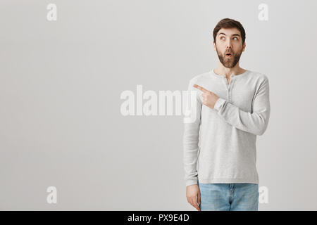 Aufgeregt verspielten europäischen Mann suchen und zeigen in der oberen linken Ecke, sagen wow und glücklich zu sein von dem, was er sieht, auf grauen Hintergrund. Fasziniert Kerl ist, etwas zu tun, lustig Stockfoto