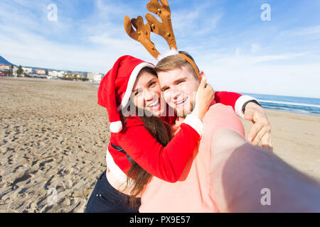Spaß und Winterurlaub Konzept - glückliches Paar in Weihnachten Kostüme unter selfie über Sandstrand Hintergrund Stockfoto