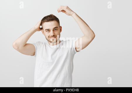 Kerl, mit coolen Frisur aus Friseur Haarschnitt berühren und lächelnd aus Freude freuen. Kunde zufrieden mit geschickten Friseur stehend über graue Wand im casual weißes T-Shirt Stockfoto