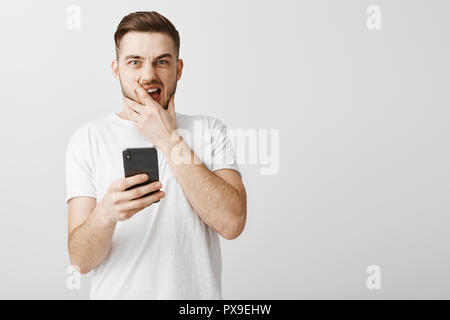 Schockiert hübscher Kerl Öffnung schreckliche Foto von ex - Freundin Schreien von Abneigung und lassen Sie sich überraschen mit geöffnetem Mund und starrte finster intensive Holding-Smartphone in der Hand betäubt Stockfoto