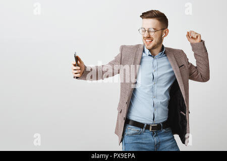 Innen- Schuss von freudiger glücklich aussehenden Europäischen männliche Unternehmer in Trendige Jacke über blaues Hemd tanzen sorglos Holding smartphone Singen Song während der Wiedergabe von Musik in drahtlosen Ohrhörer Stockfoto