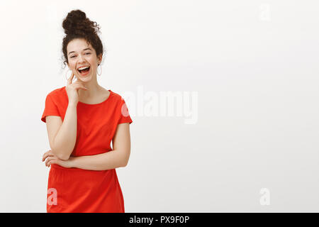 Portrait von Sorglos glücklich europäische Mädchen in trendigen rotes Kleid mit lockigem Haar von Bun gekämmt, laughing out loud und berühren mit der Hand, mit Freunden zu reden, sich in City Cafe über graue Wand Stockfoto