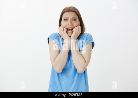 Portrait von verängstigten europäische Frau mit blondem Haar, schreiend und runzelte die Stirn und Hände halten in der Nähe der geöffneten Mund, weil sie Angst haben und entsetzt, Schreck, zitternd vor Angst über graue Wand Stockfoto