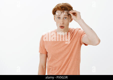 Portrait von charmanten nerdy rothaarige Kerl mit Sommersprossen in orange T-Shirt, Brille und Blick mit Erstaunen und Überraschung an Kamera, beeindruckt zu sehen, unglaublich charmanten Mitschüler Stockfoto