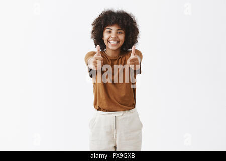 Ja sagen zu neuen Möglichkeiten. Hell und fröhlich afrikanische amerikanische Mädchen mit Afro Frisur im stilvollen braunes T-Shirt ziehen die Hände in Richtung Kamera mit Daumen nach oben und lächelnd zujubeln und unterstützende Stockfoto