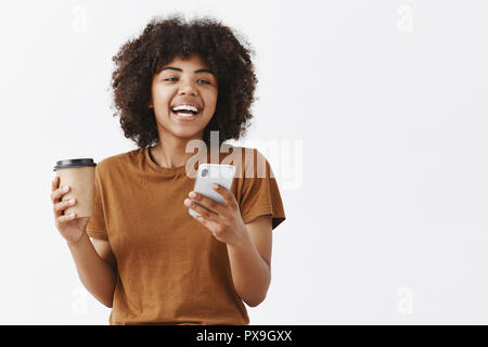 Taille-up Shot von trendigen Sorglos afrikanische amerikanische Mädchen mit lockigem Haar in Braun t-shirt Lachen beim Gespräch mit Freunden trinken Kaffee aus dem Pappbecher und halten über grauer Hintergrund smartphone Stockfoto