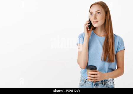 Studio geschossen von Verträumten gut aussehende Frau mit Ingwer Haar und Sommersprossen links mit nostalgischen Blick schauen, die eine neue Tasse Kaffee während Anrufen Freund holding Smartphone in der Nähe der Ohr Stockfoto