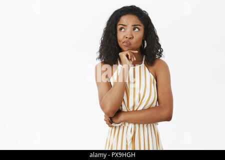 Unsicher, intelligente und kreative attraktive african american Female in trendigen gestreiften gelbe Overalls grinsend die Hand am Kinn in nachdenklicher Pose Blick auf die obere linke Ecke, denken Wahl Stockfoto