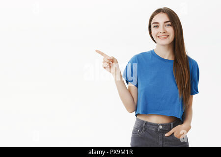 Freundlich ausgehende Schöne junge Brünette in blauen T-Shirt die Hand in der Tasche der Jeans links und lächelt nett und höflich an Kamera, als ob zeigt Weg zu Kunden oder Förderung etwas Stockfoto