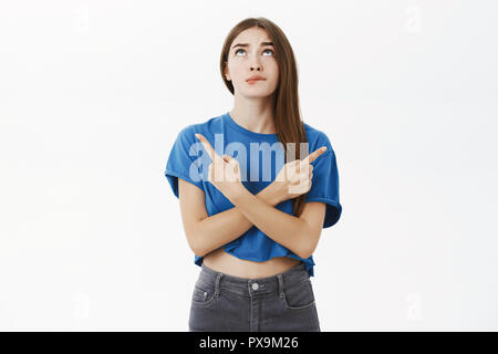 Hmm Mädchen Zögern bei der Wahl. Portrait von Unsicher wunderschöne Frauen in blauen T-Shirt beißen Lippen, blickte unsicher Kreuzung Hände gegen den Körper nach links und rechts über poinding graue Wand Stockfoto