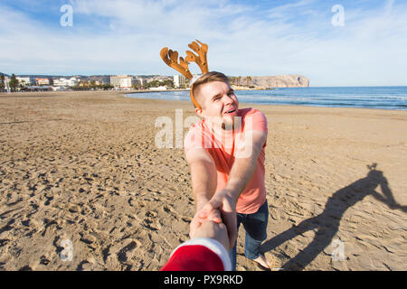 Folgen Sie mir, Ferien und Weihnachten Konzept-fröhlicher junger Mann mit Hirsch Hörner holding Santa's Hand am Strand. Stockfoto