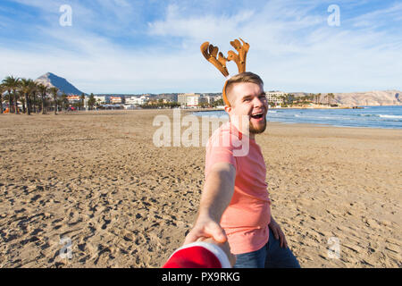 Folgen Sie mir, Ferien und Weihnachten Konzept-fröhlicher junger Mann mit Hirsch Hörner holding Santa's Hand am Strand. Stockfoto