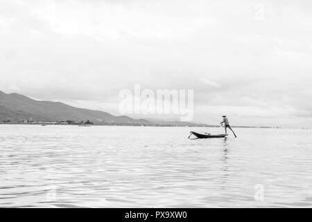 Reisen lokale junge burmesen männliche Fischer tragen geprüft, t-shirt, mit Stick und net zu fischen, Balancieren auf einem Fuß auf dem Boot, Inle Lake Myanmar, Birma Stockfoto