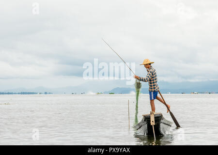 Reisen lokale junge burmesen männliche Fischer tragen geprüft, t-shirt, mit Stick und net zu fischen, Balancieren auf einem Fuß auf dem Boot, Inle Lake Myanmar, Birma Stockfoto