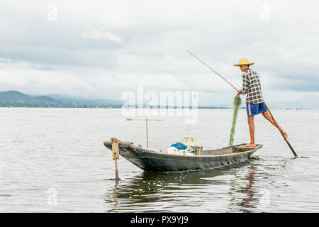 Reisen lokale junge burmesen männliche Fischer tragen geprüft, t-shirt, mit Stick und net zu fischen, Balancieren auf einem Fuß auf dem Boot, Inle Lake Myanmar, Birma Stockfoto