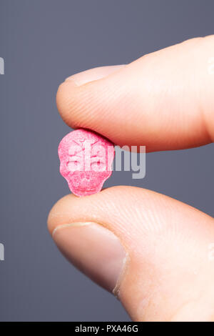 Nahaufnahme der kaukasischen männlichen Finger halten ein wenig rosa XTC, MDMA oder Medikamente Pille auf einem grauen Hintergrund. Stockfoto
