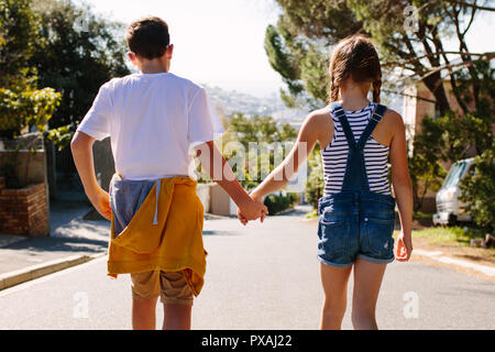 Kinder in der Liebe zu Fuß auf einer Straße, die Hand in Hand an einem sonnigen Tag. Junge und Mädchen zu Fuß auf einer verlassenen Straße zusammen. Stockfoto