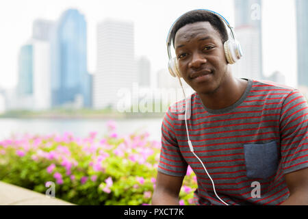 Junge schöne afrikanischer Mann im Park sitzen beim Hören von Musik über Kopfhörer Stockfoto