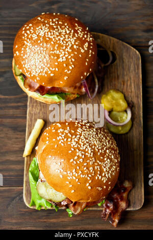 Hamburger mit gebratenem Speck, Zwiebeln, Salat und marinierten Gurken auf Holzbrett. Ansicht von oben, flach Stockfoto